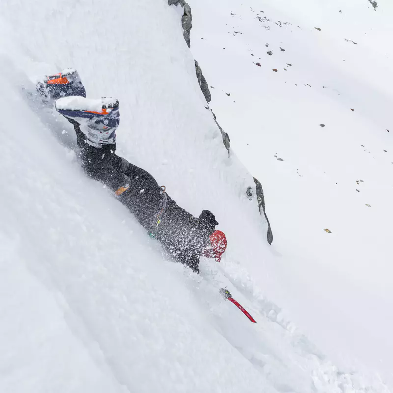 Een persoon in winterkleding en met een helm valt van een steile, besneeuwde helling af, waarbij de sneeuw omhoog spuit terwijl hij naar beneden glijdt. Op de achtergrond zijn rotsen en ongerepte sneeuw te zien.